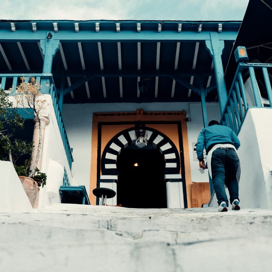 Escalier bleu menant à une porte voûtée, une silhouette y grimpant, sous un ciel couvert